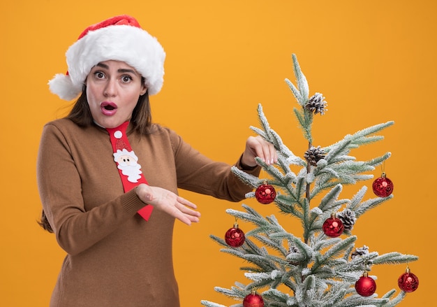 surprised young beautiful girl wearing christmas hat with tie standing nearby christmas tree isolated on orange wall