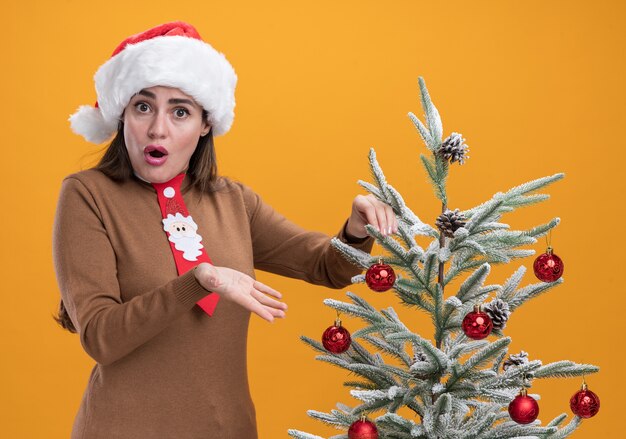 オレンジ色の壁に隔離されたクリスマス ツリーの近くに立っているネクタイでクリスマス帽子をかぶっている美しい少女を驚かせた
