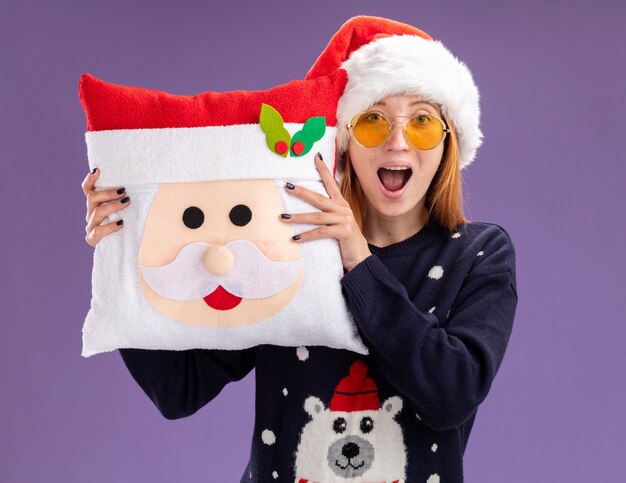 Surprised young beautiful girl wearing christmas sweater and hat with glasses holding christmas pillow isolated on purple background