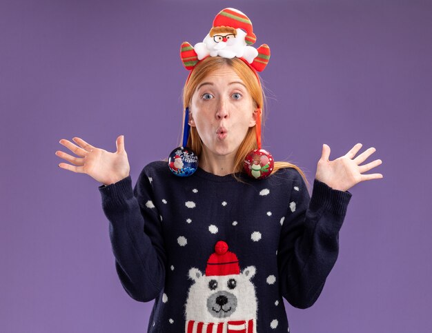 Удивленная молодая красивая девушка в рождественском свитере с рождественским обручем для волос повесила елочные шары на ухе, разводя руками, изолированными на фиолетовом фоне