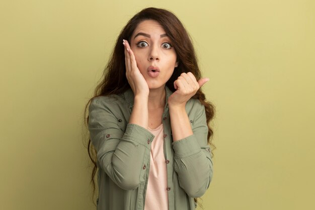 Surprised young beautiful girl wearing olive green t-shirt putting hand on cheek and points at side isolated on olive green wall with copy space