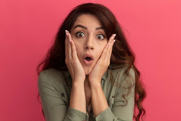 Surprised young beautiful girl wearing olive green t-shirt putting hands on cheek isolated on pink wall