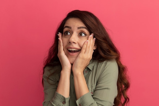 Surprised young beautiful girl wearing olive green t-shirt putting hands on cheeks isolated on pink wall