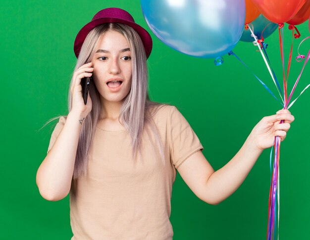 Surprised young beautiful girl wearing party hat and braces holding balloons speaks on phone 