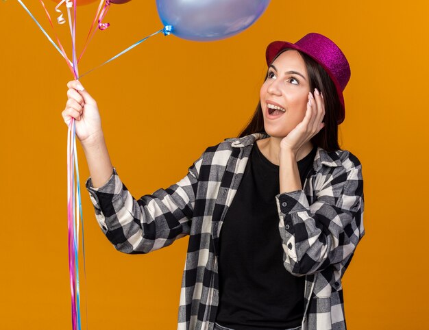 Surprised young beautiful girl wearing party hat holding balloons putting hand on cheek isolated on orange wall