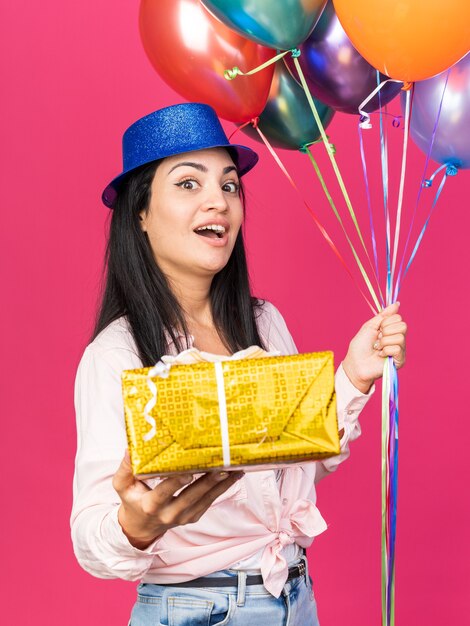 Surprised young beautiful girl wearing party hat holding balloons with gift box isolated on pink wall