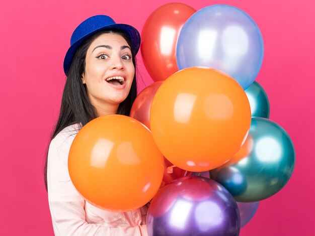 Surprised young beautiful girl wearing party hat holding balloons 