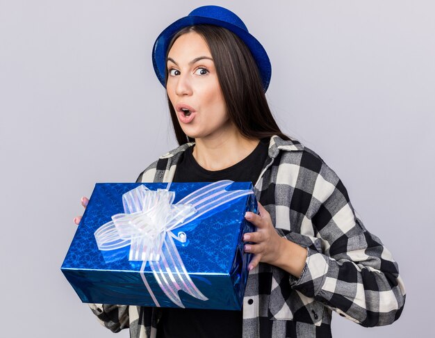 Surprised young beautiful girl wearing party hat holding gift box 