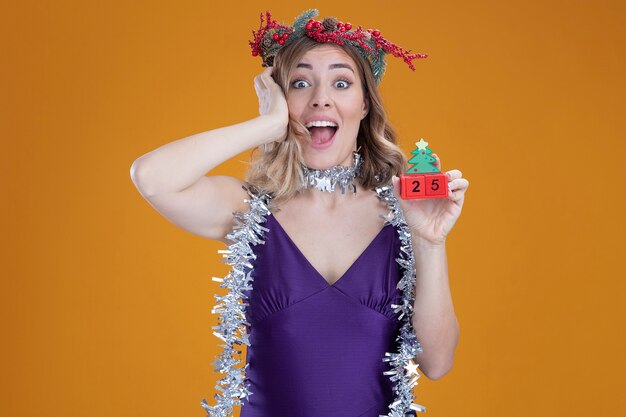 茶色の背景で隔離の頭に手を置いてクリスマスのおもちゃを保持している首に花輪と紫色のドレスと花輪を身に着けている驚いた若い美しい少女