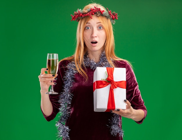 Surprised young beautiful girl wearing red dress with wreath and garland on neck holding glass of champagne with gift box isolated on green background