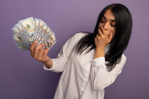 Surprised young beautiful girl wearing white t-shirt holding and looking at cash isolated on purple