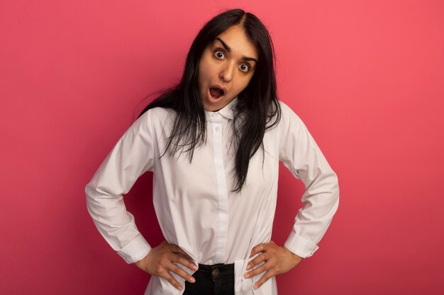 Surprised young beautiful girl wearing white t-shirt putting hands on hip isolated on pink