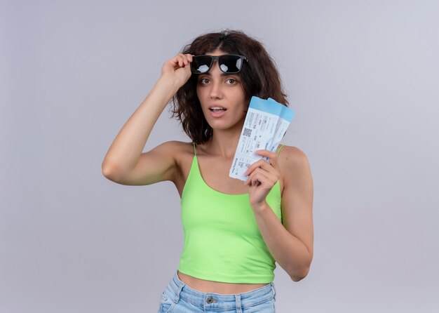 Surprised young beautiful traveler woman wearing and holding sunglasses and holding airplane tickets on isolated white wall