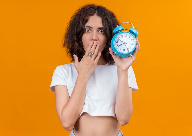 Surprised young beautiful woman holding alarm clock and putting hand on mouth on isolated orange wall with copy space