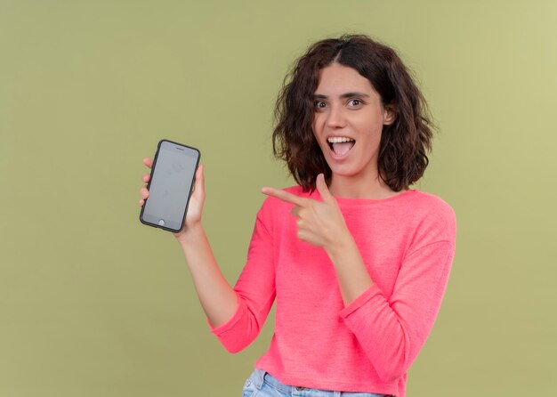 Surprised young beautiful woman holding mobile phone and pointing at it on isolated green wall