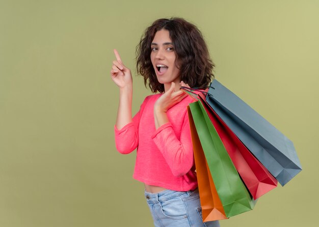 Surprised young beautiful woman holding paper bags and pointing up on isolated green wall with copy space