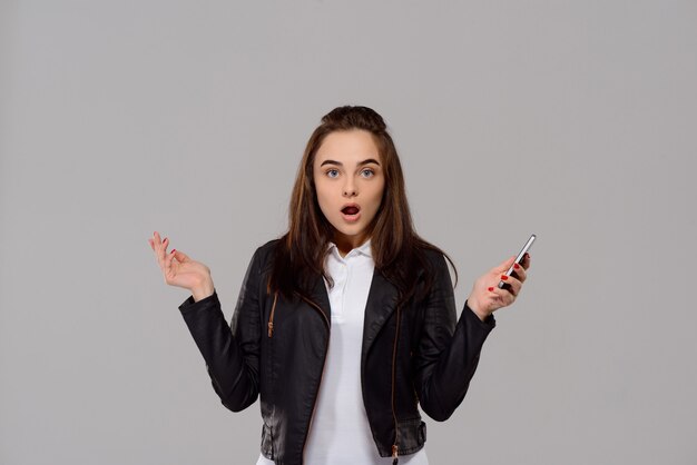 Surprised young beautiful woman holding phone over purple wall