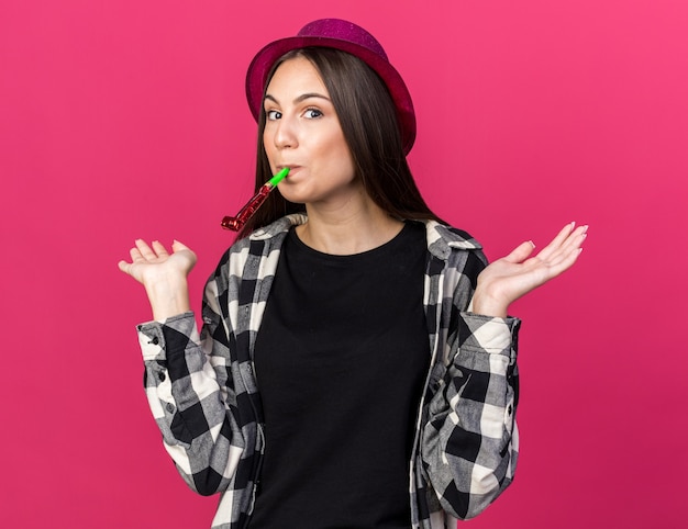 Surprised young beautiful woman wearing party hat blowing party whistle spreading hands isolated on pink wall