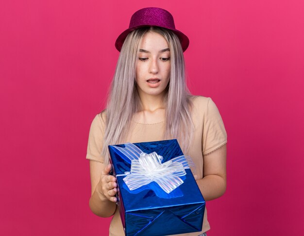 Surprised young beautiful woman wearing party hat holding and looking at gift box isolated on pink wall