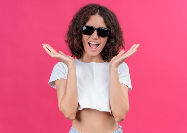 Surprised young beautiful woman wearing sunglasses and showing empty hands on isolated pink wall with copy space