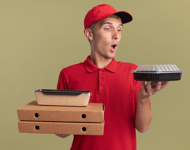 놀란 금발 배달 소년은 피자 상자에 음식 패키지를 들고 복사 공간이 있는 올리브 녹색 벽에 격리된 식품 용기를 봅니다.