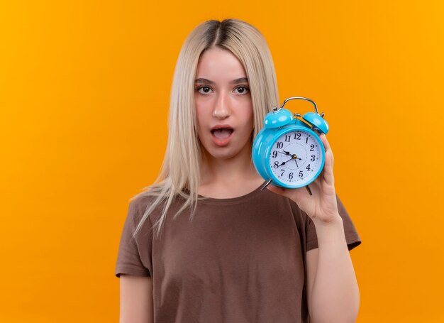 Surprised young blonde girl holding alarm clock on isolated orange wall with copy space