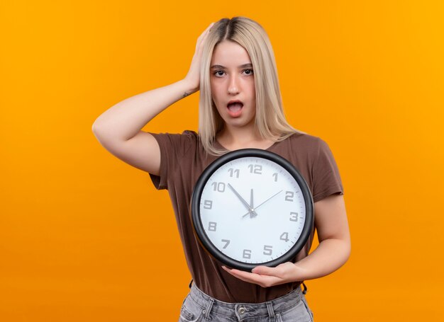 Surprised young blonde girl holding clock with hand on head on isolated orange wall with copy space