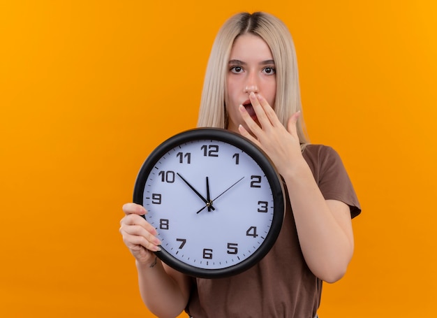 Surprised young blonde girl holding clock with hand on mouth on isolated orange wall with copy space