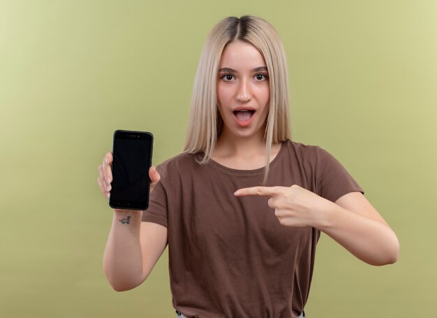 휴대 전화를 들고 고립 된 녹색 벽에 그것을 가리키는 놀란 된 젊은 금발 소녀