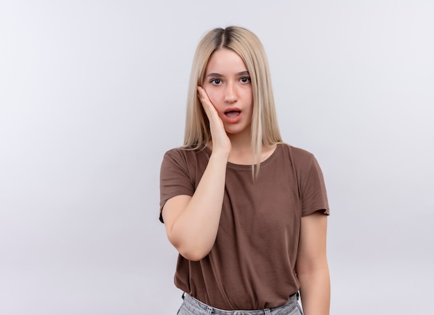 Surprised young blonde girl putting hand on cheek on isolated white wall with copy space