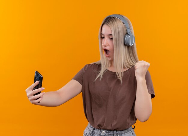 Удивленная молодая блондинка в наушниках держит мобильный телефон, глядя на него с поднятым кулаком на изолированной оранжевой стене