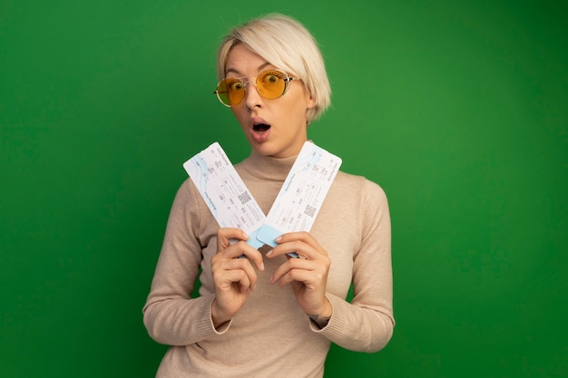 Foto gratuita giovane ragazza bionda sorpresa che indossa gli occhiali da sole che tengono i biglietti dell'aereo isolati sulla parete verde con lo spazio della copia