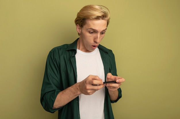 Удивленный молодой блондин в зеленой футболке играет по телефону
