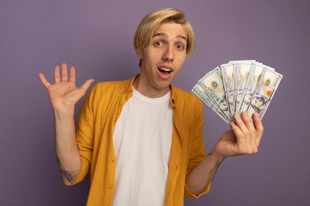 Free photo surprised young blonde guy wearing yellow t-shirt holding cash and raising hand isolated on purple