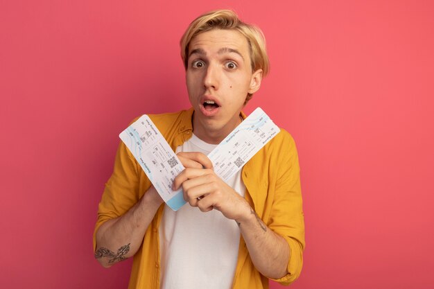 Surprised young blonde guy wearing yellow t-shirt holding tickets isolated on pink