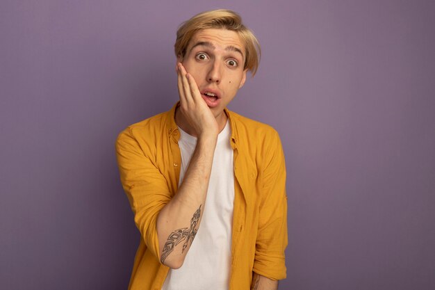 Surprised young blonde guy wearing yellow t-shirt putting hand on cheek isolated on purple