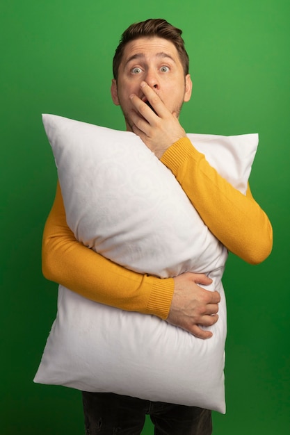 Surprised young blonde handsome man hugging pillow looking straight putting hand on mouth isolated on green wall