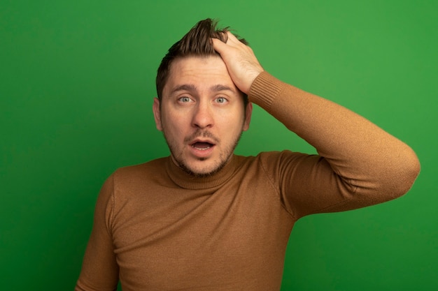 Free photo surprised young blonde handsome man putting hand on head looking at front isolated on green wall