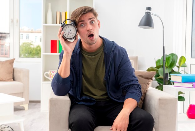 Surprised young blonde handsome man sits on armchair holding alarm clock