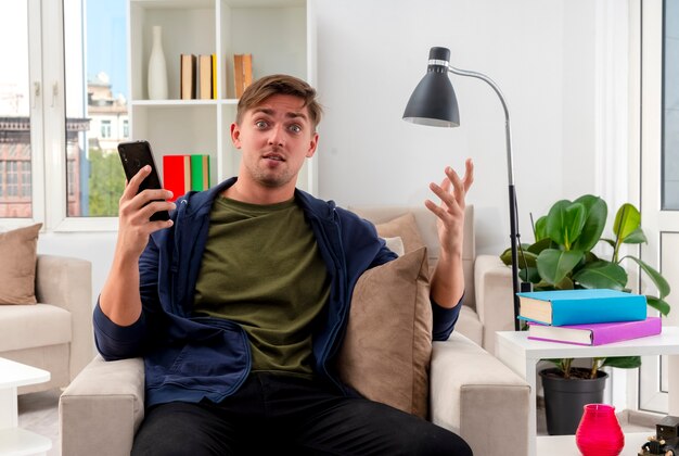 Surprised young blonde handsome man sits on armchair holding phone and raising hand inside the living room