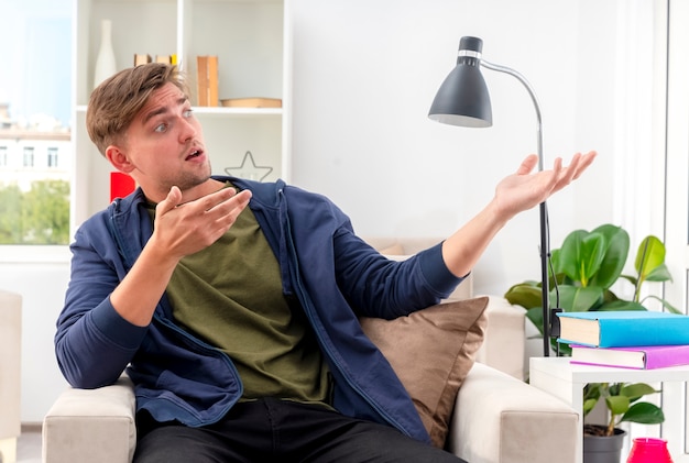 Surprised young blonde handsome man sits on armchair looking and pointing at side with two hands inside the living room
