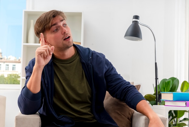 Surprised young blonde handsome man sits on armchair points up looking at side inside the living room