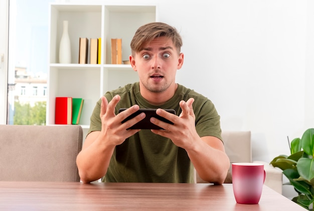 Surprised young blonde handsome man sits at table with cup holding and looking at phone inside the living room