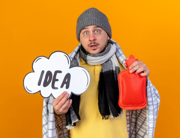 Удивленный молодой блондин, больной славянский мужчина в зимней шапке и шарфе, завернутый в плед, держит пузырь идеи и бутылку с горячей водой, изолированные на оранжевой стене с копией пространства