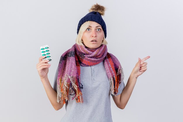 Surprised young blonde ill slavic woman wearing winter hat and scarf holds pack of medical pills and points at side isolated on white wall with copy space