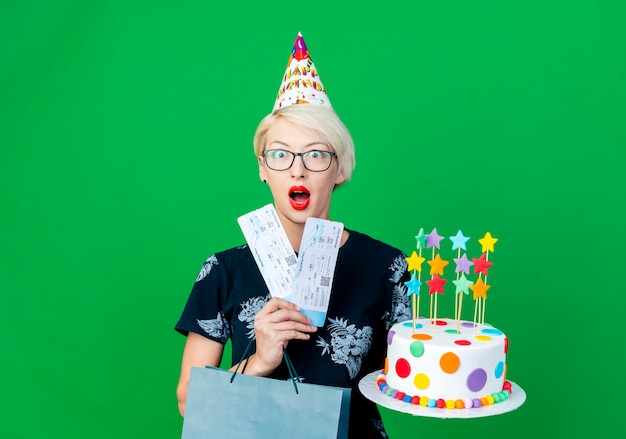 Surprised young blonde party girl wearing glasses and birthday cap holding birthday cake with stars airplane tickets and paper bag looking at camera isolated on green background with copy space