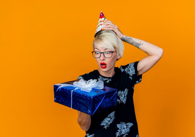 Surprised young blonde party girl wearing glasses and birthday cap holding and looking at gift box keeping hand on cap isolated on orange background with copy space