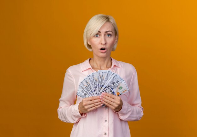 Surprised young blonde slavic woman holding cash isolated on orange