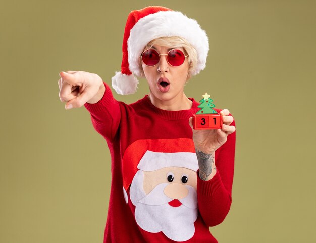 surprised young blonde woman wearing christmas hat and santa claus christmas sweater with glasses holding christmas tree toy with date looking and pointing at side isolated on olive green wall with copy space