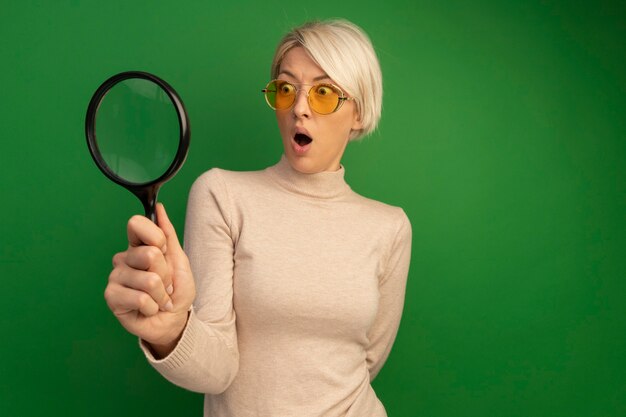 Surprised young blonde woman wearing sunglasses holding magnifying glass looking at side through it keeping hand behind back isolated on green wall with copy space